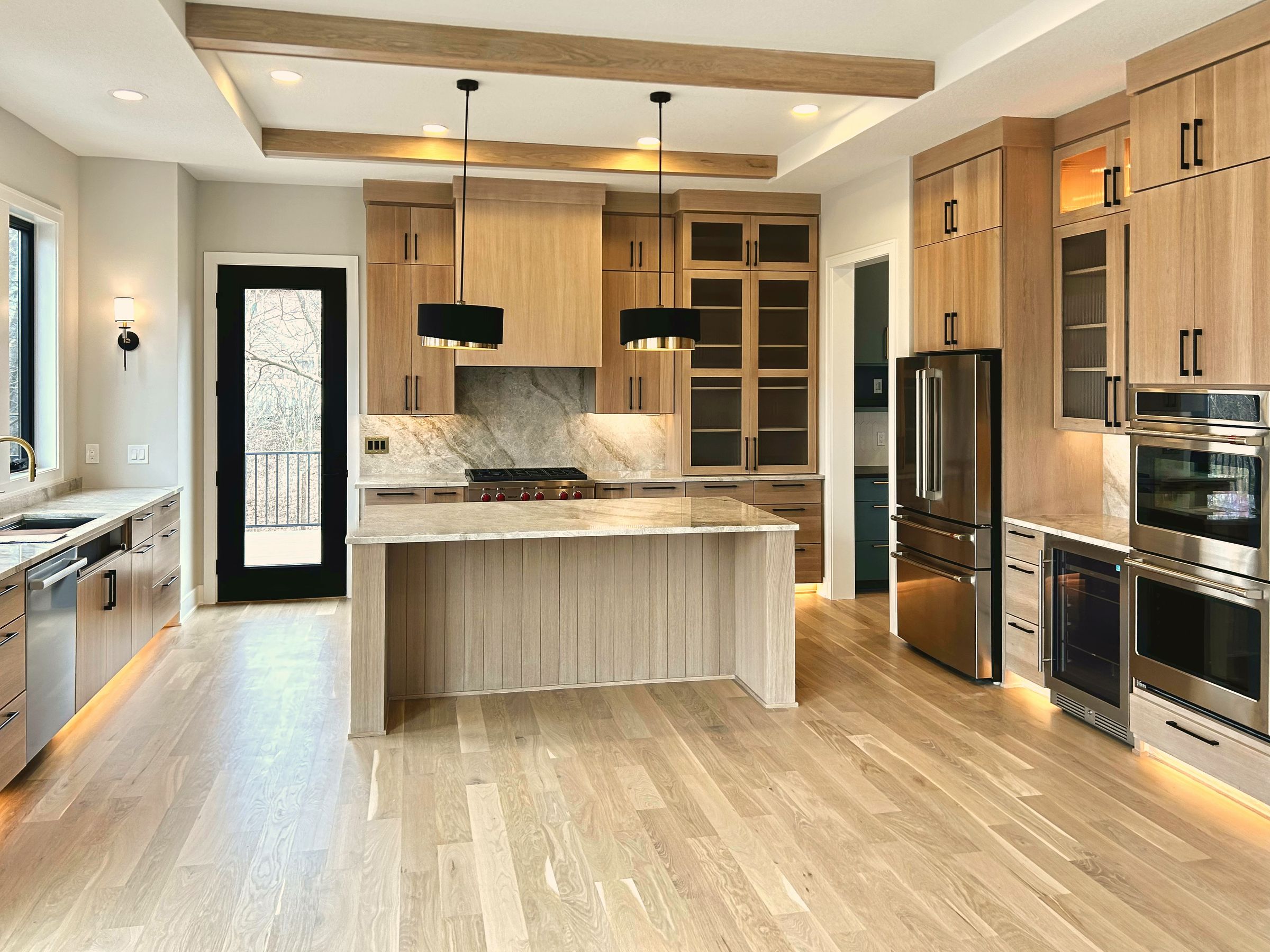 Kitchen with rift white oak slab cabinetry, a stone backsplash and island, black drum pendants, and stained ceiling beams
