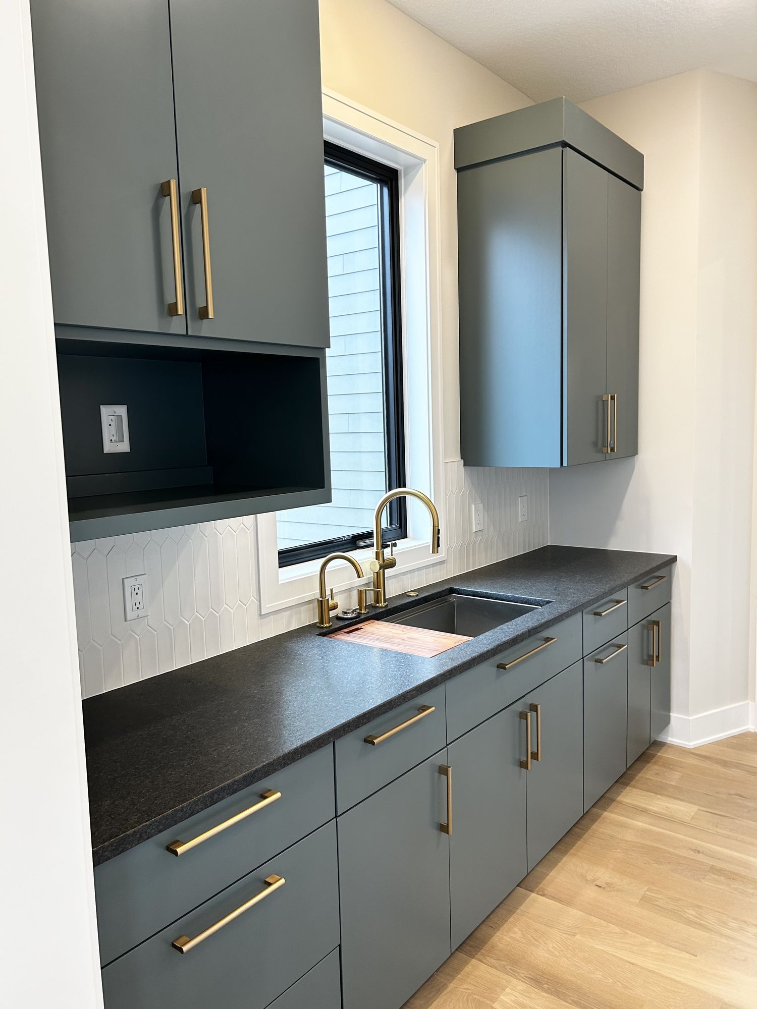Butler's pantry with green-gray slab cabinetry, brass pulls and faucet, a black stone countertop, and a white picket tile backsplash
