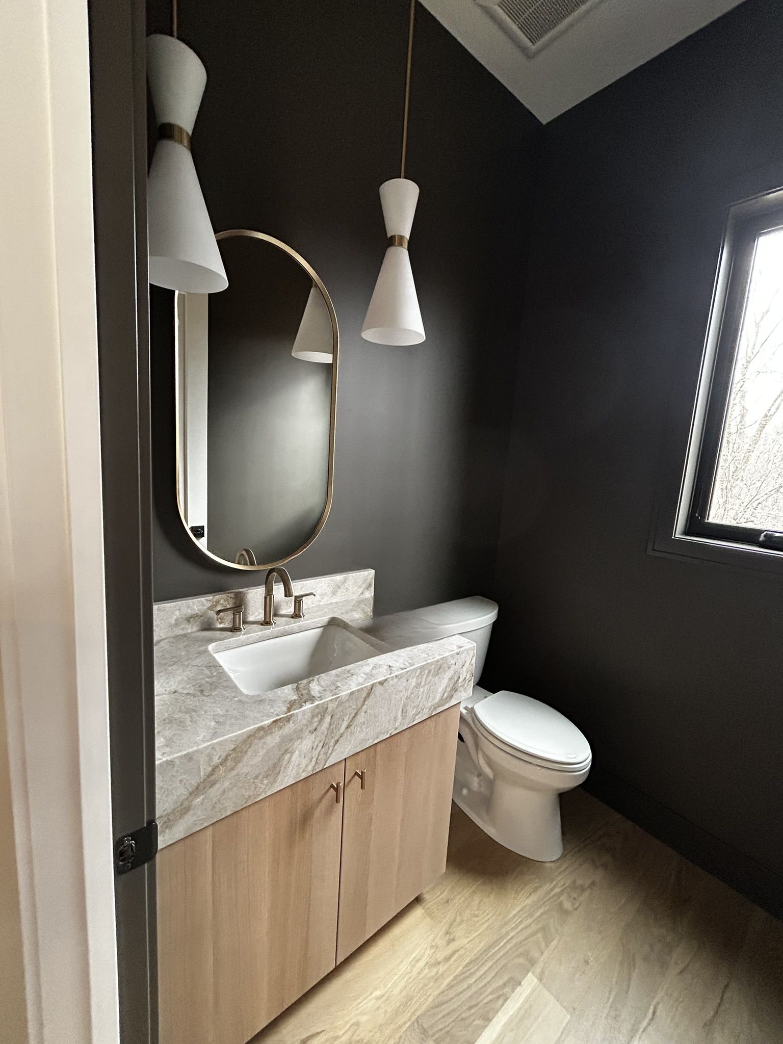 Powder bath with charcoal walls, a white oak vanity under a thick stone top, an oval brass mirror, and white cone pendants