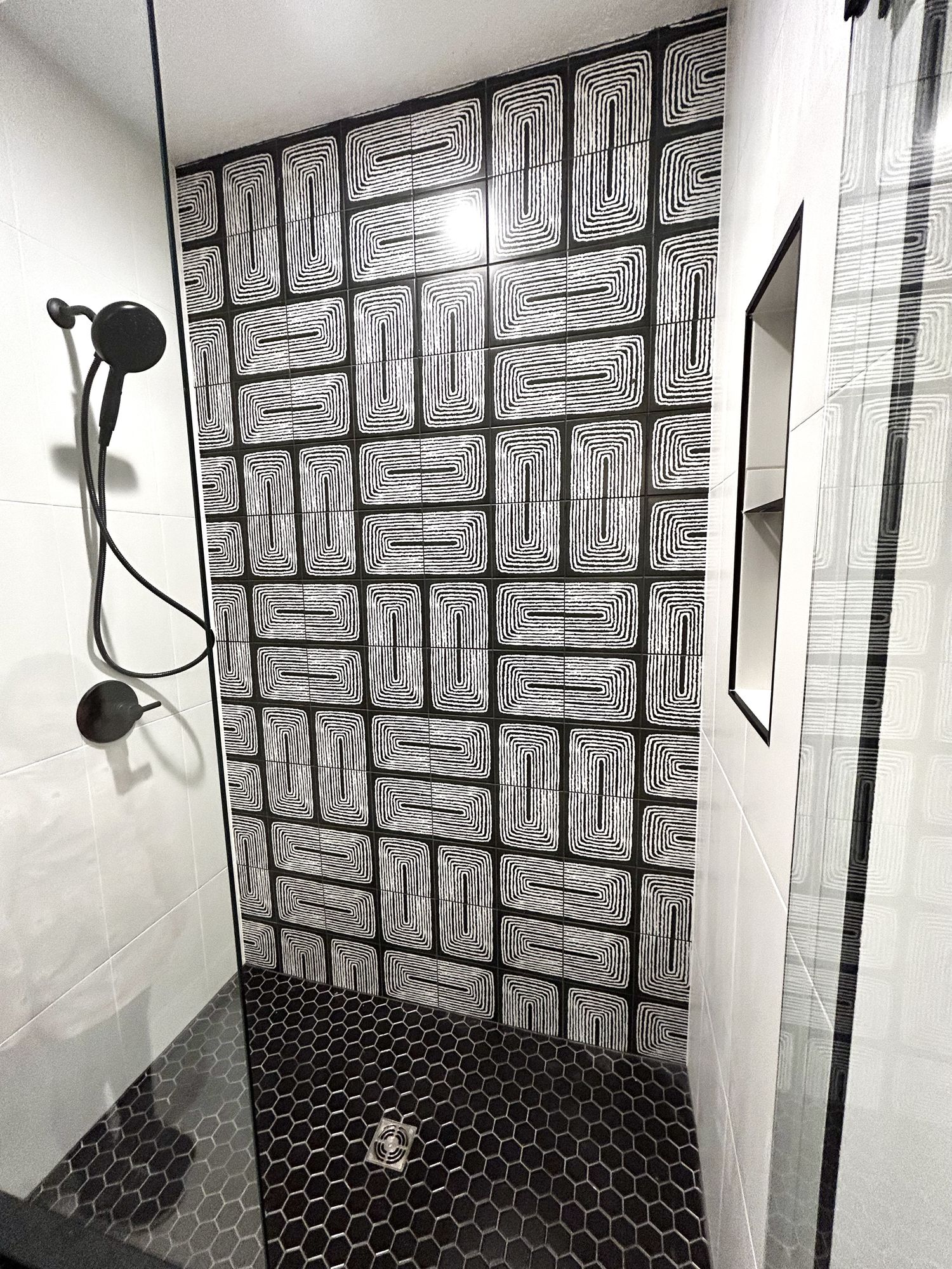 Shower with a black-and-white graphic tile wall, a black hex mosaic floor, and matte black fixtures
