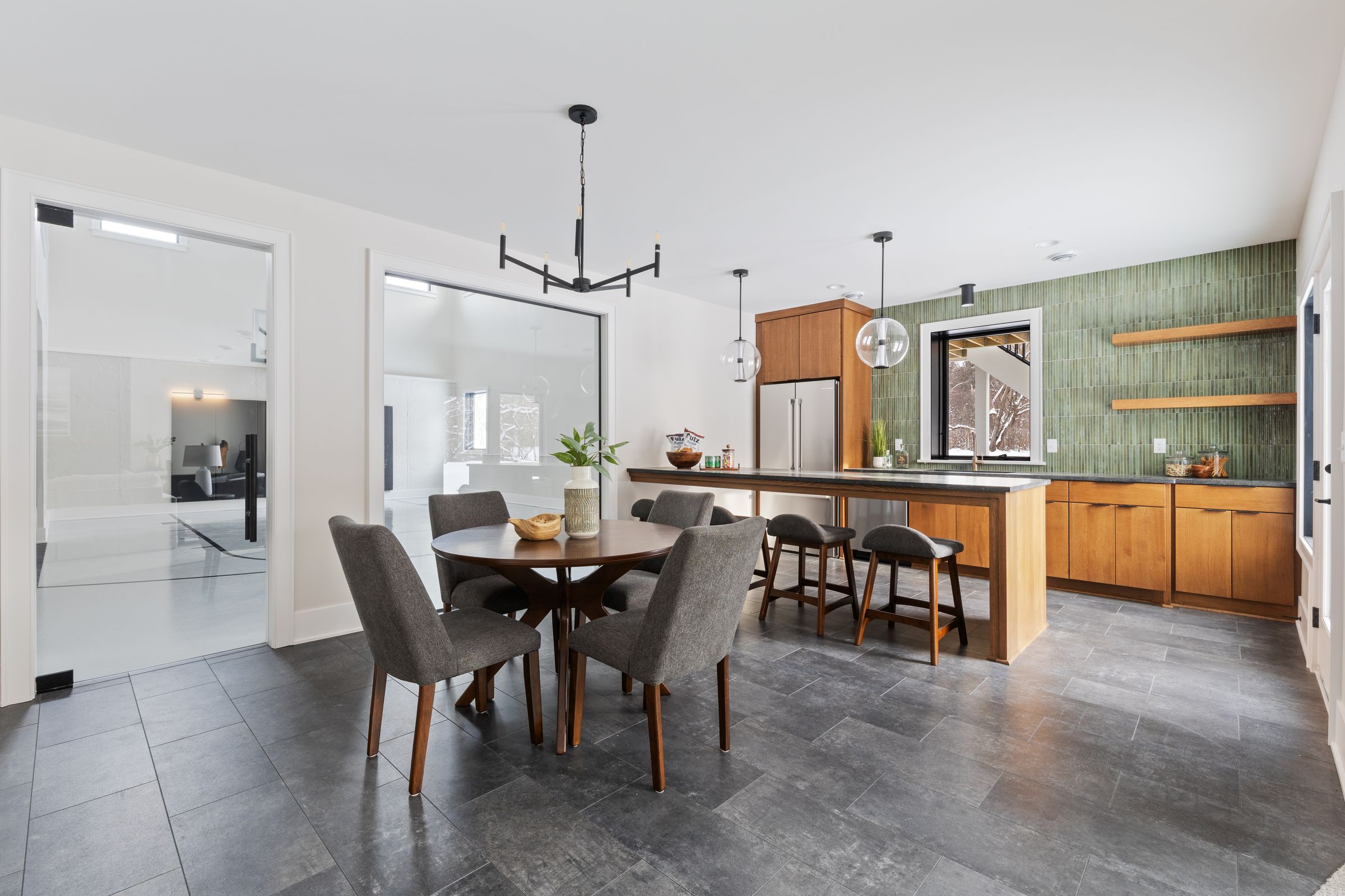 Lower-level bar with green vertical stacked tile, warm wood slab cabinetry and floating shelves, a dark stone counter, glass globe pendants, and a dining table beside glass doors