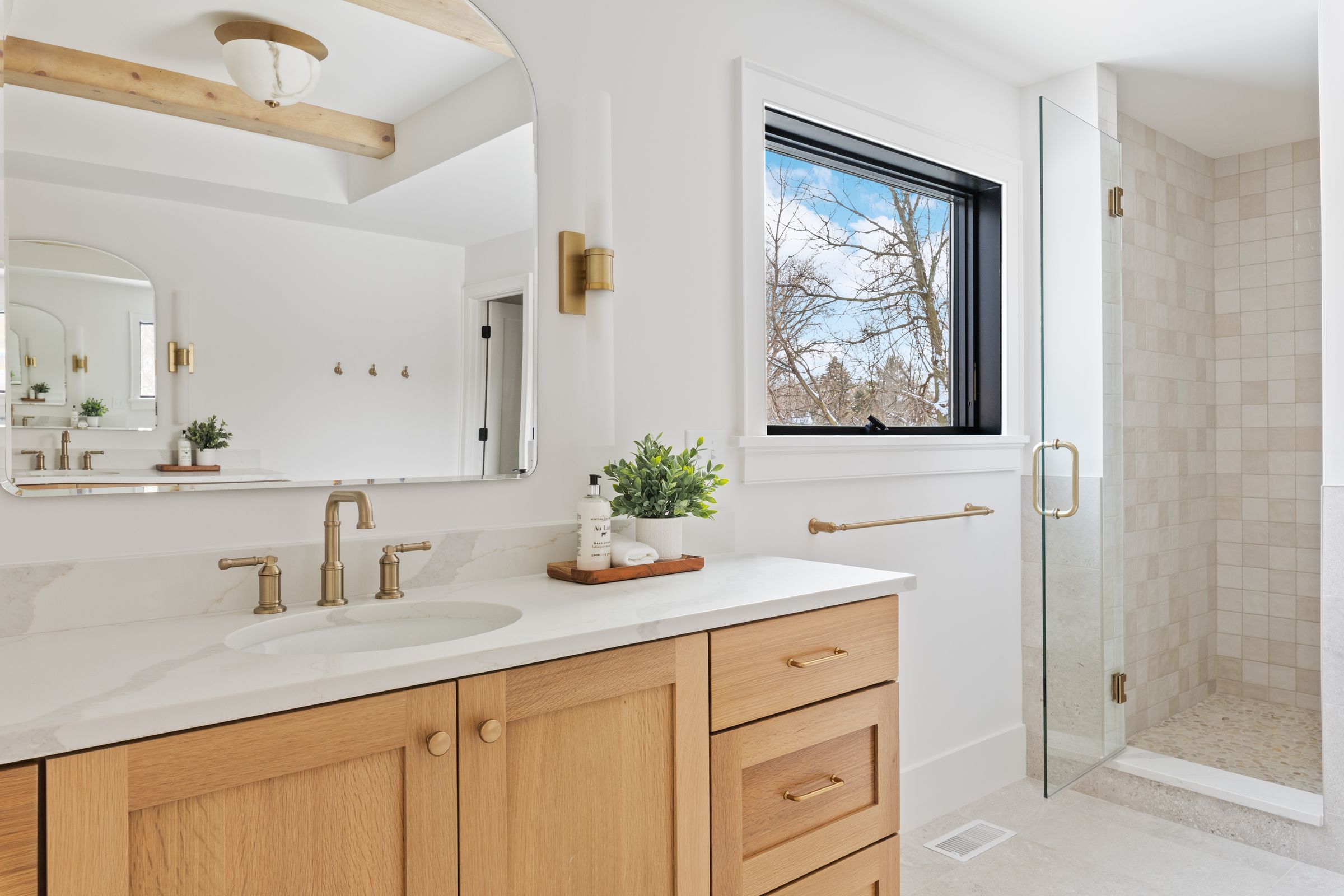 Primary bath with a white oak vanity, brass fixtures, an arched mirror, and a walk-in shower with square tile