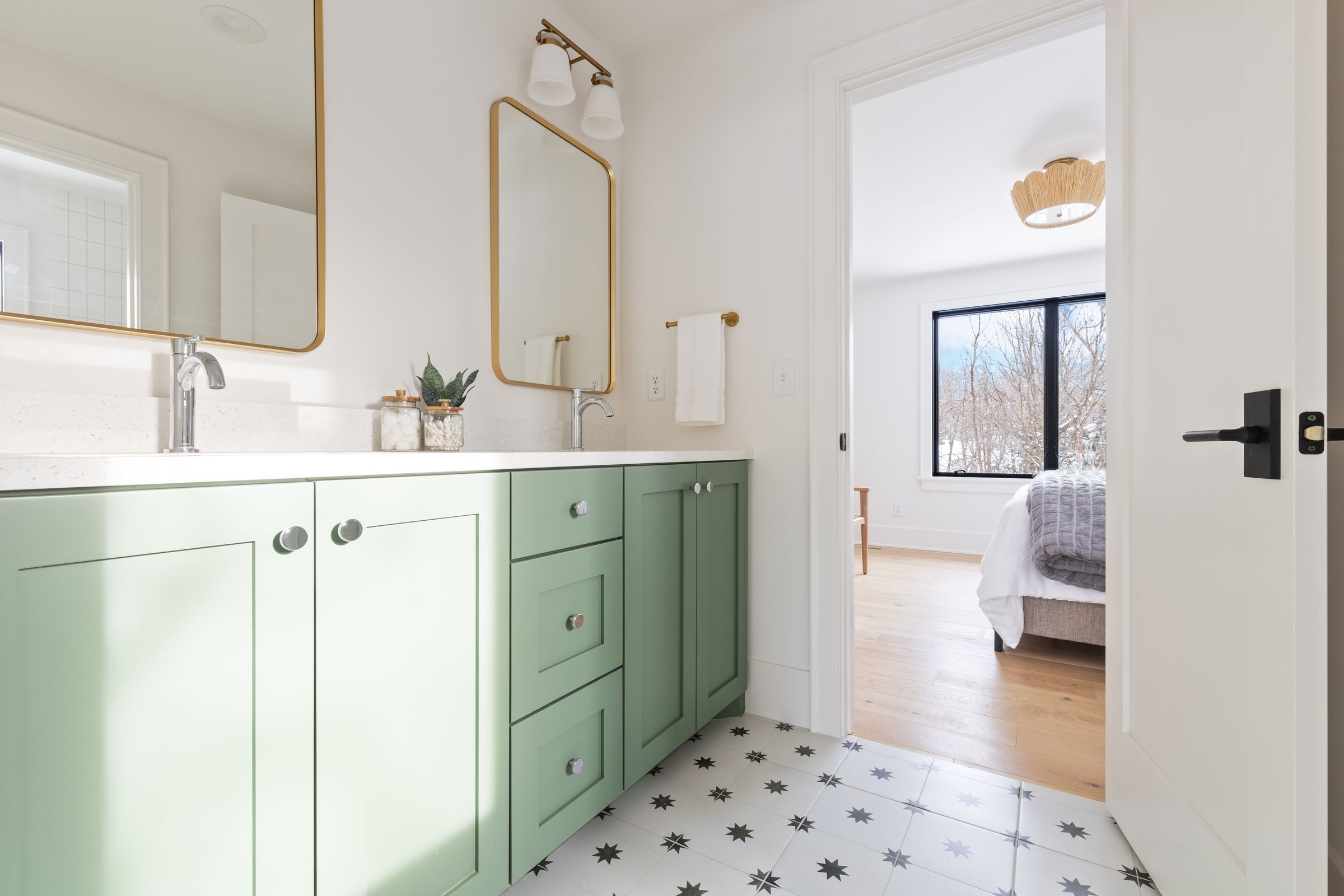 Secondary bath with a sage green double vanity, brass-framed mirrors and sconce, a black and white star tile floor, and a view into the bedroom