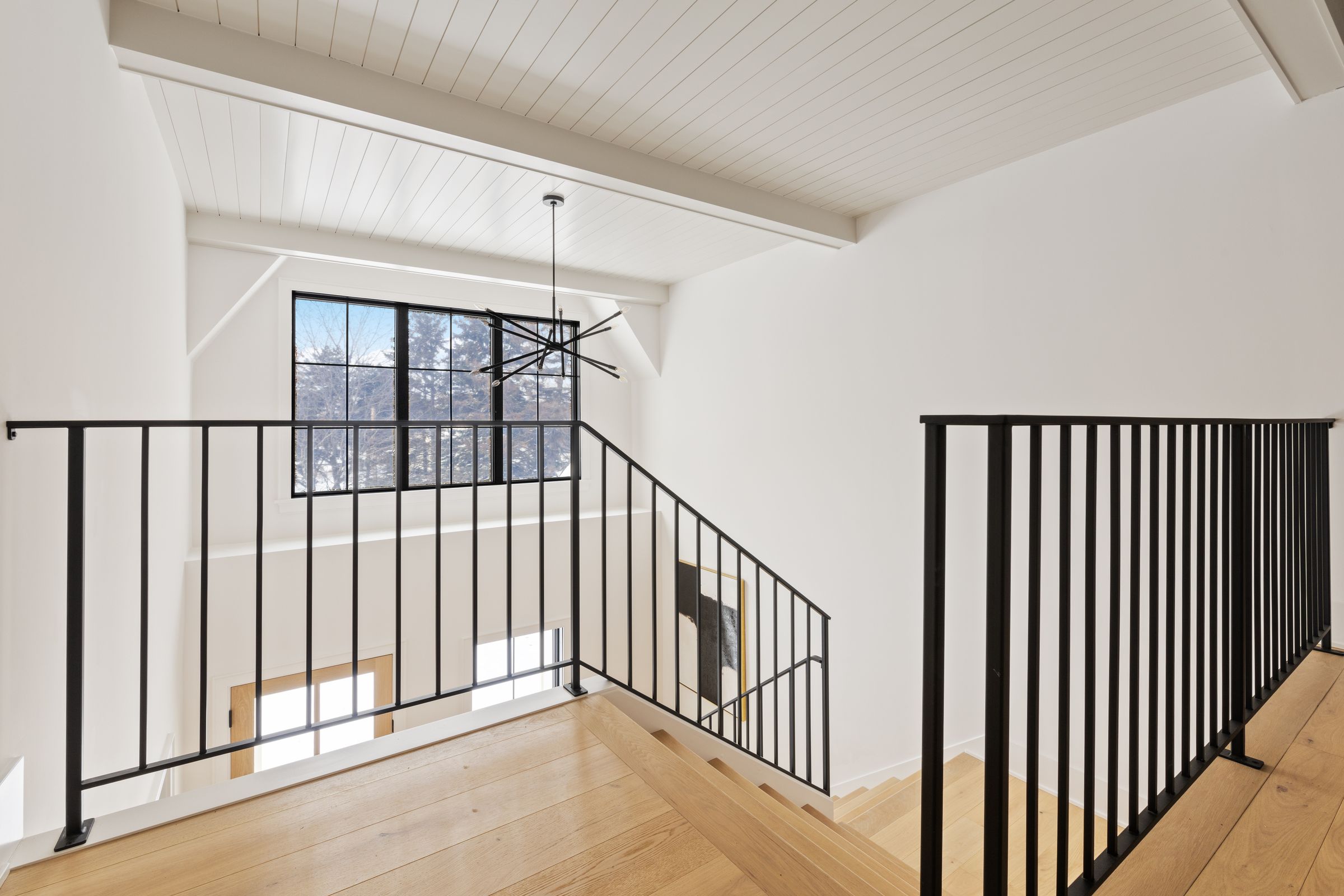 Upper stair landing with black metal railings, a white plank ceiling, and a sculptural black chandelier