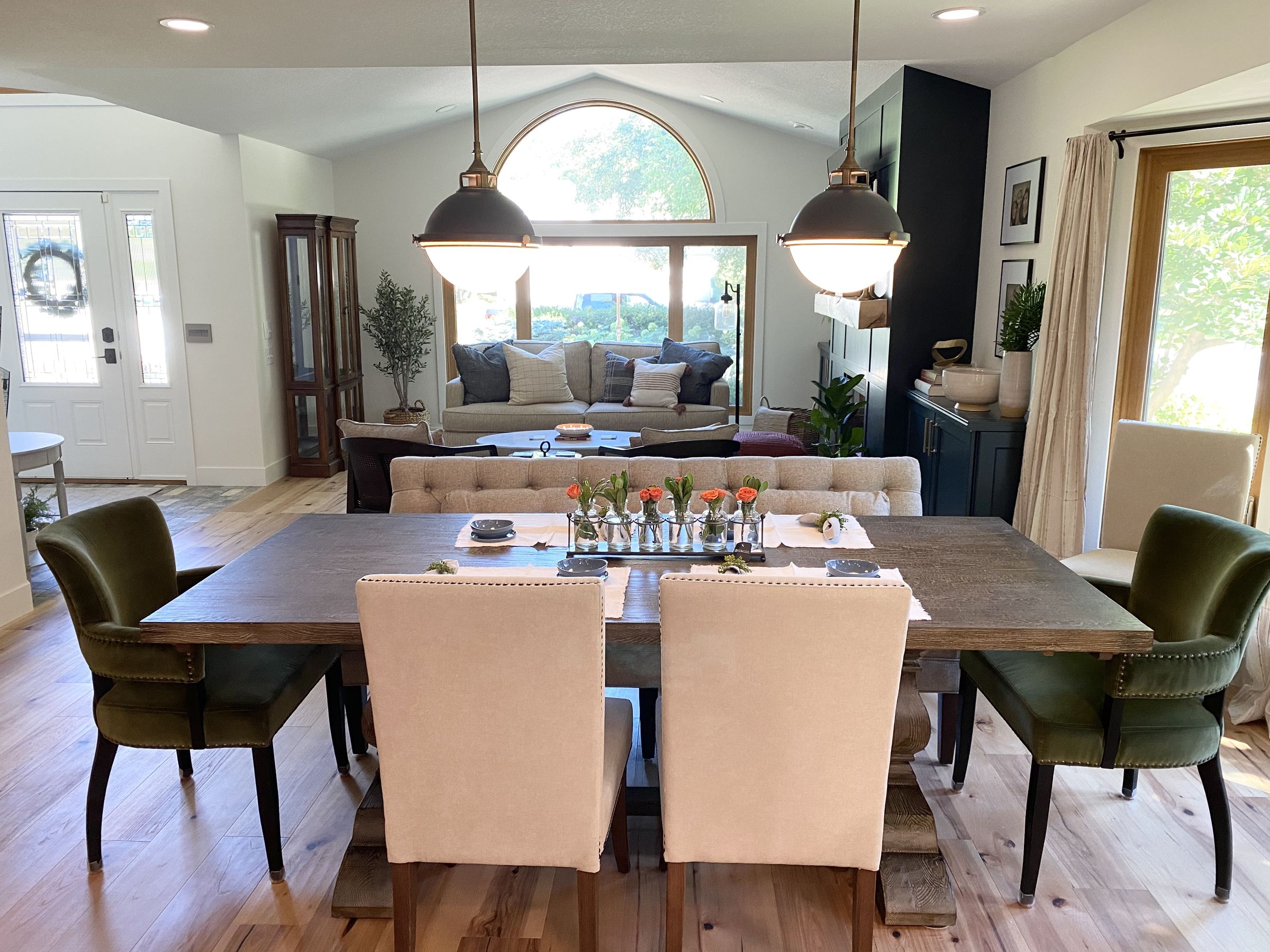 Dining table with green velvet and linen chairs under two globe pendants, looking past a tufted settee into the living room with an arched window