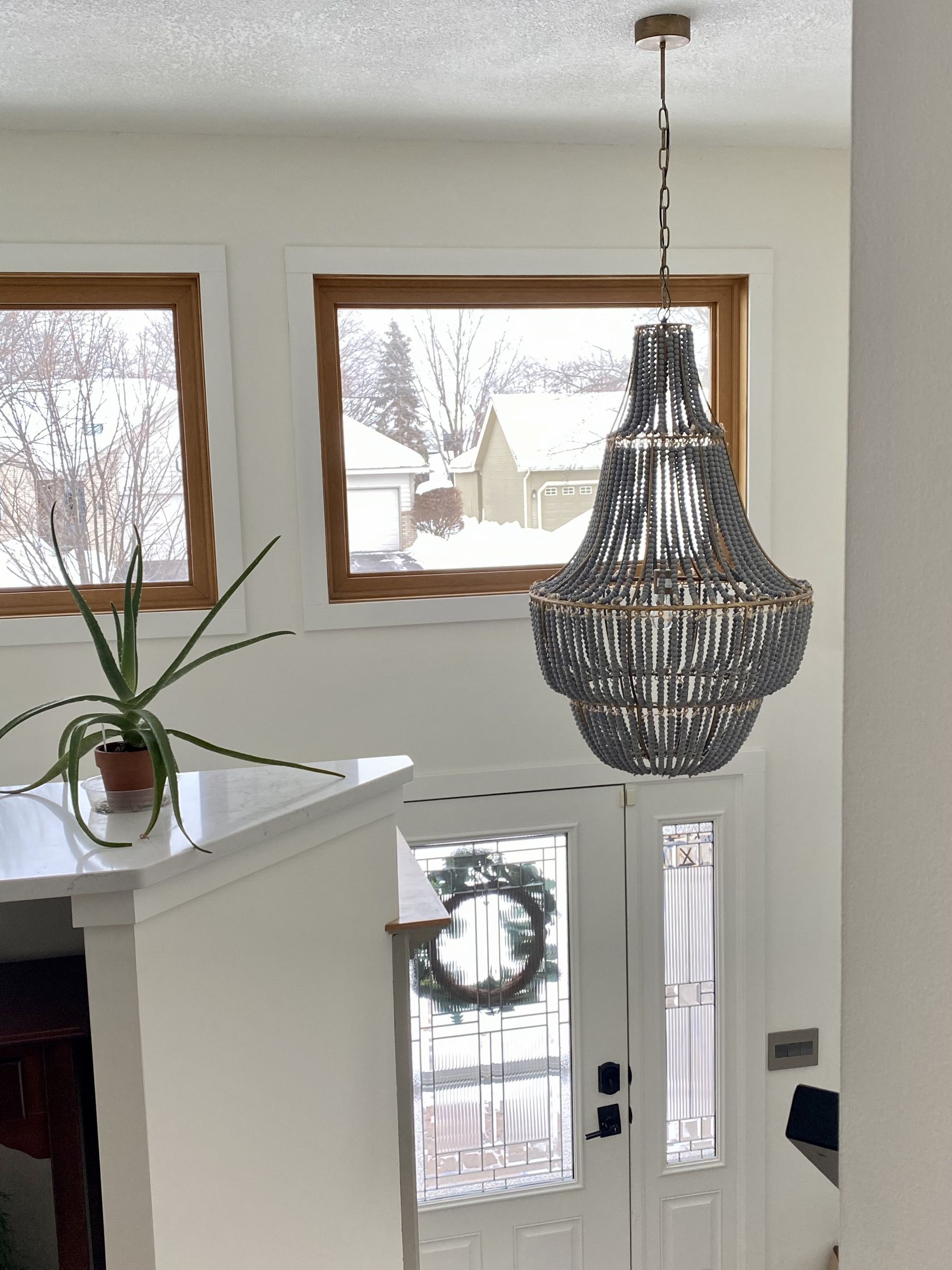 Gray beaded chandelier hanging over the split-level entry, with white walls, wood-trimmed windows, and a glass-paneled front door