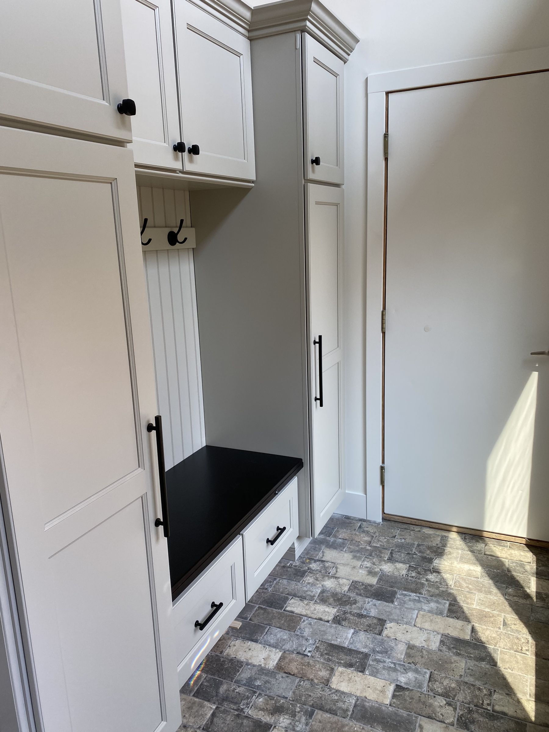Custom entry locker cabinet in pale gray with a beadboard back, hooks, a black bench top, and drawers below, over a brick-look tile floor