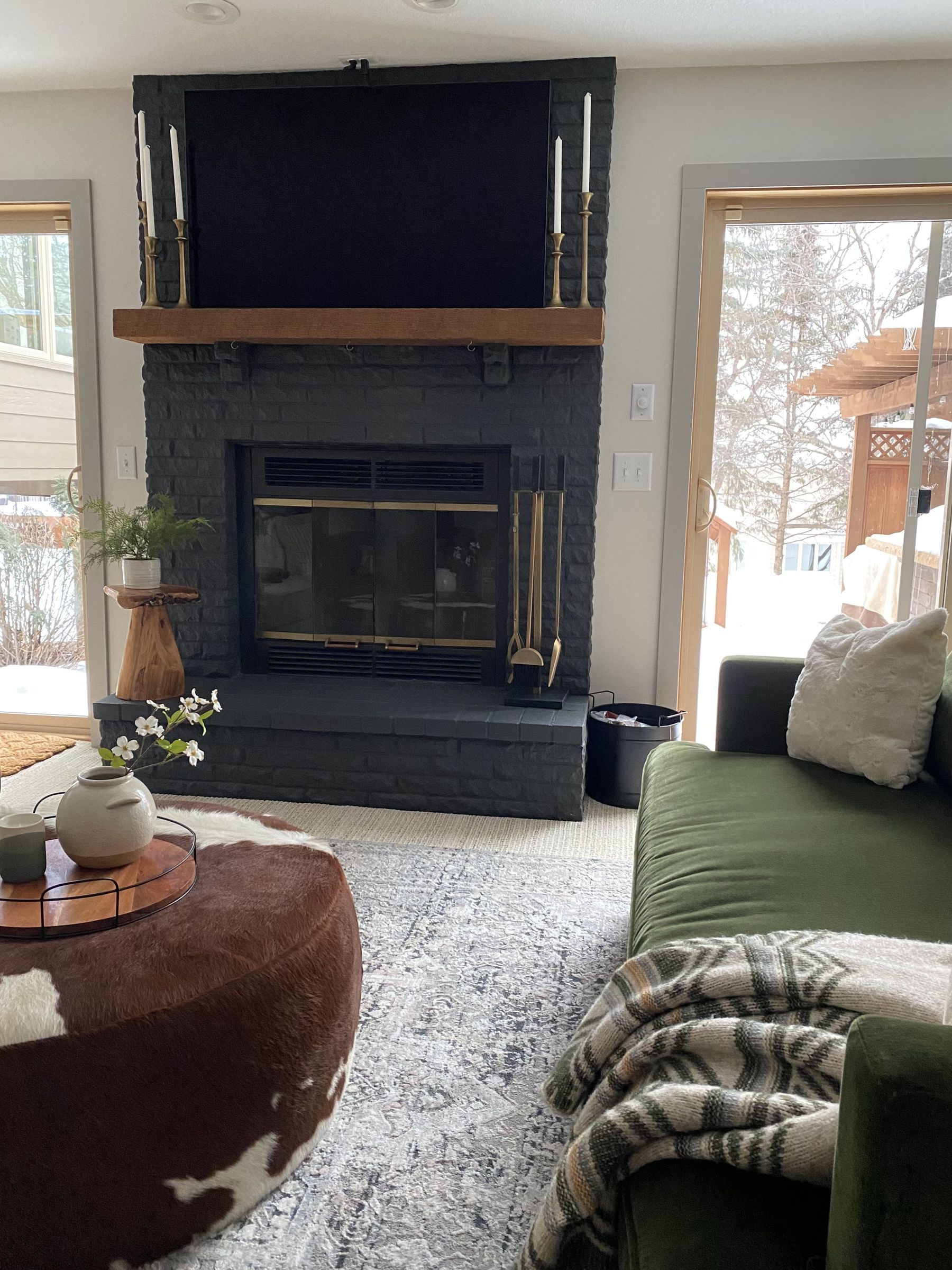 Family room, furnished and decorated, with a charcoal painted brick fireplace, a green velvet sofa, and a cowhide ottoman