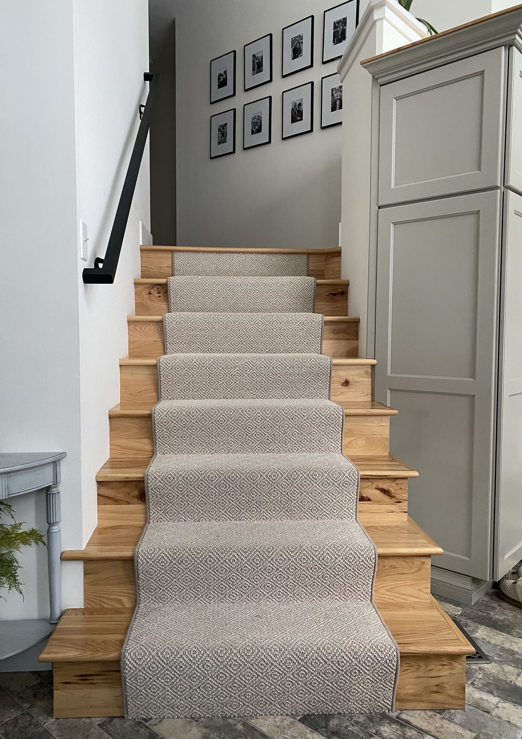 Hickory stairs with a patterned runner, a black metal handrail, and a gallery wall of black-and-white family photos