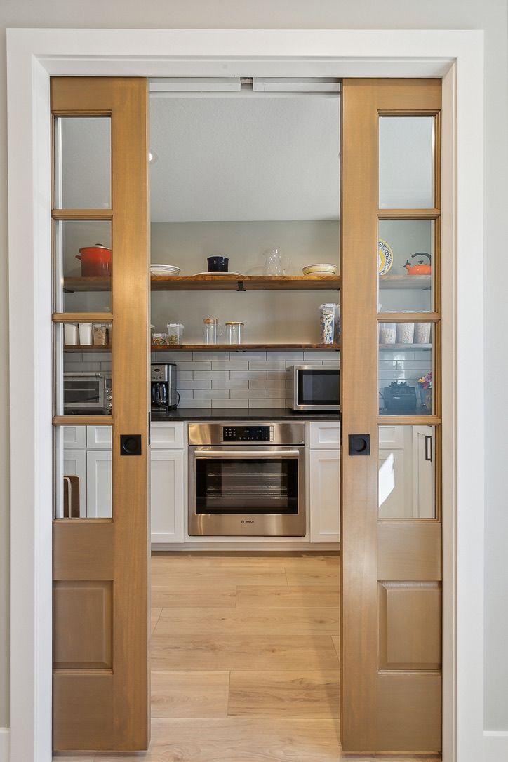 Walk-in pantry behind wood pocket doors with glass panes, with open wood shelving, a white subway tile wall, and a built-in wall oven