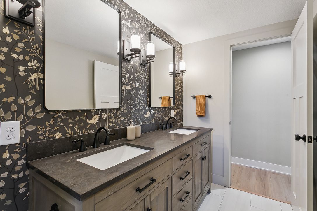 Bathroom with a dark botanical wallpaper, a stained double vanity with a dark stone top, black fixtures, and two framed mirrors with sconces