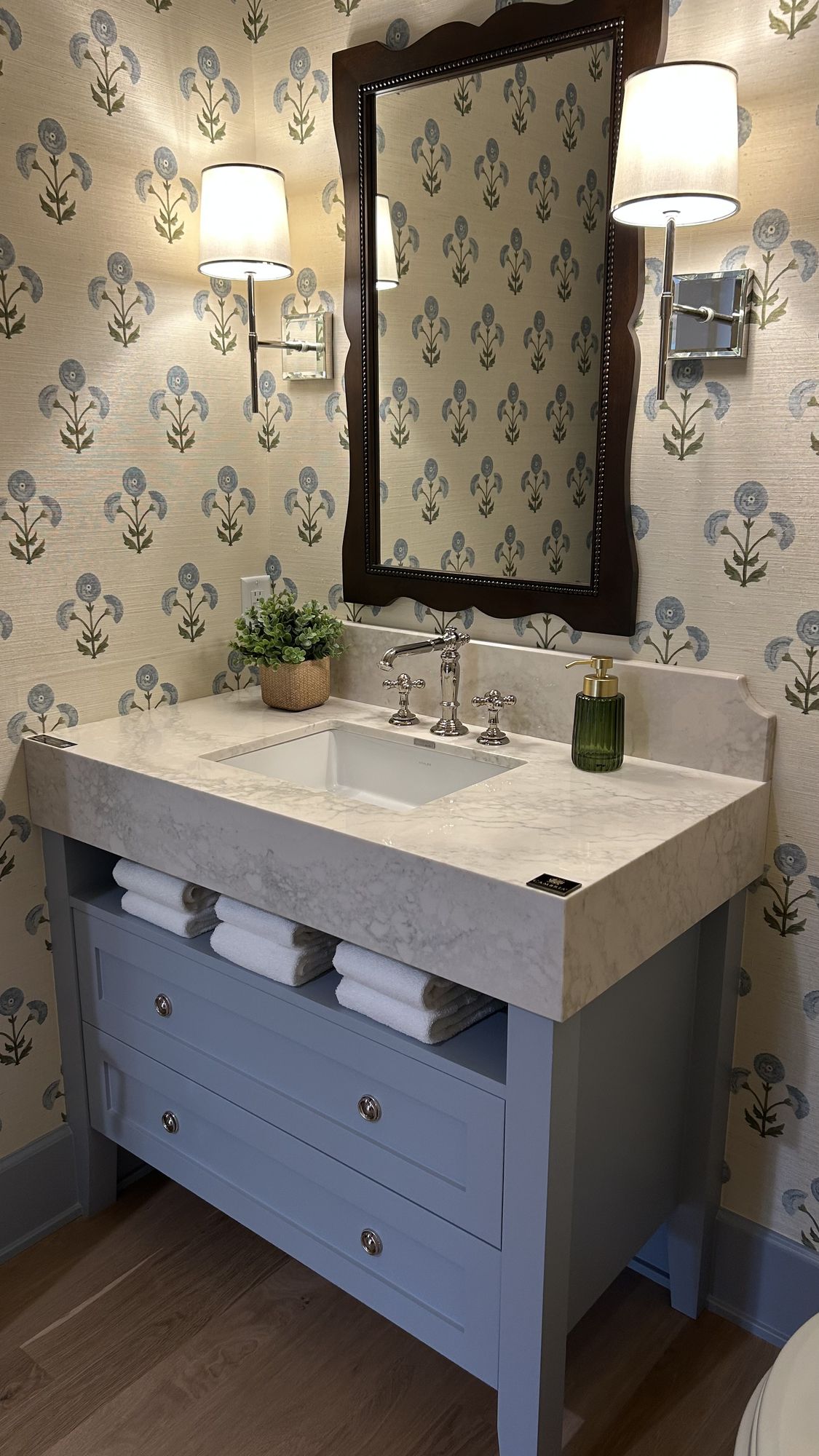 Powder bath with floral grasscloth wallpaper, a blue-gray furniture-style vanity with an open towel shelf, and a scalloped wood mirror between two sconces