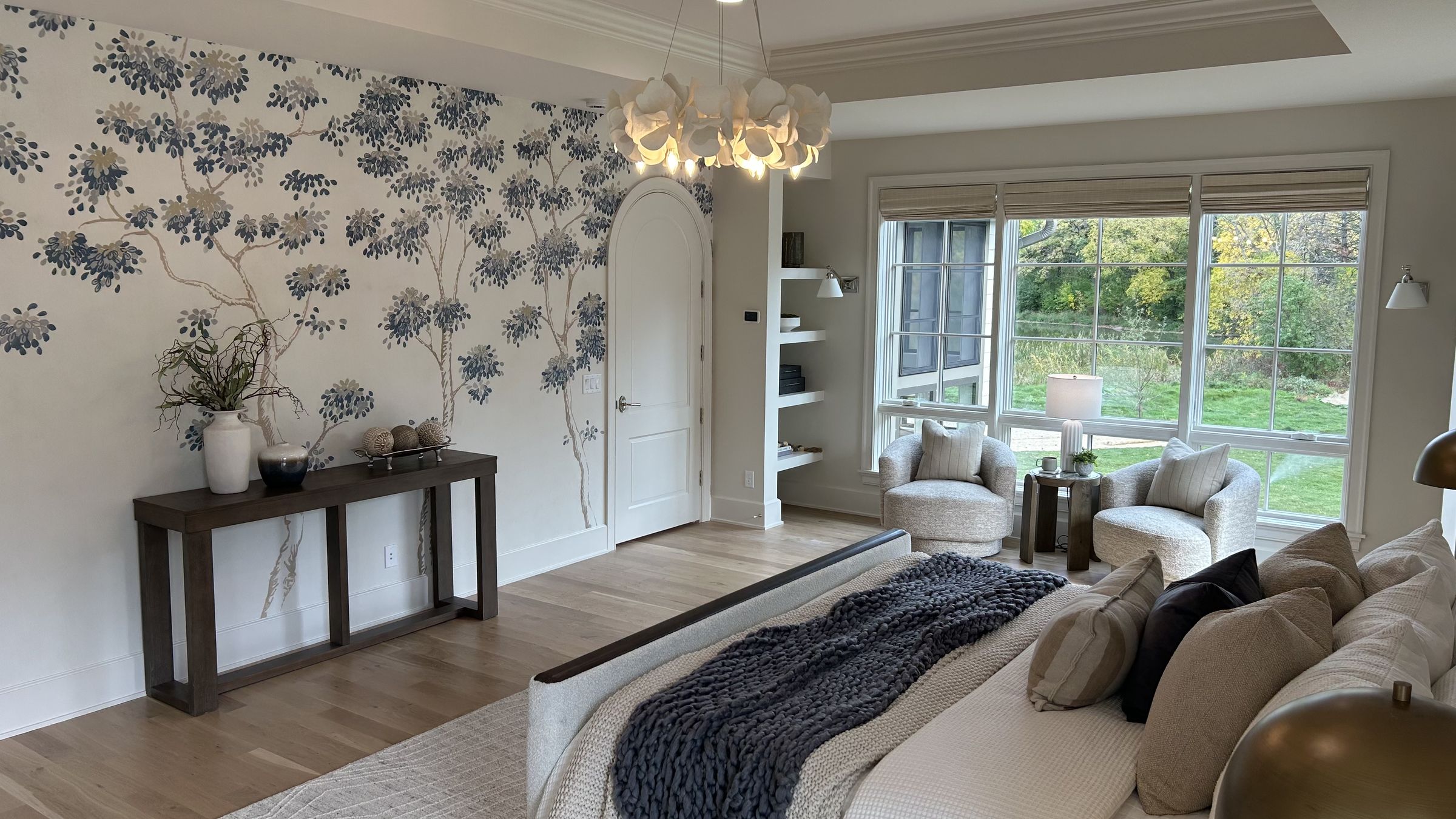 Primary bedroom with blue tree mural wallpaper, an arched door, a sculptural petal chandelier, built-in shelves, and two swivel chairs by a wall of windows